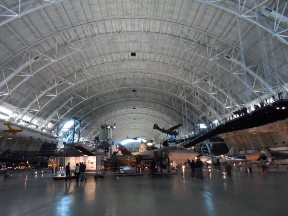 Interior of a large aircraft hangar with multiple planes on display and people walking around.