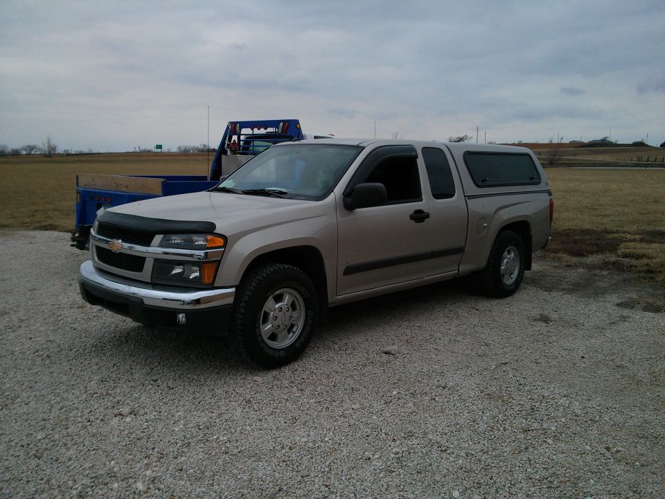 Tan Chevrolet Colorado pickup truck with a camper shell parked on gravel. Another blue truck is behind it