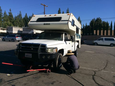 1997 Dodge 3500 - A person works on the front tire of a white Dodge Ram pickup truck fitted with a camper shell, which is supported by jacks.