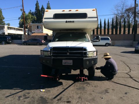 1997 Dodge 3500 - A person in a hat works on the front wheel of a Dodge Ram truck fitted with a camper, which is supported by jacks.