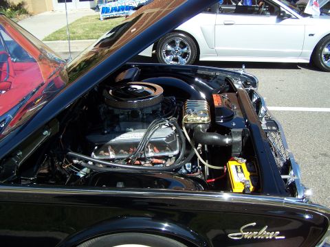 1971 Ford Ranchero - The engine bay of a black classic car is open, revealing its mechanical components. A modern silver convertible is parked behind it.