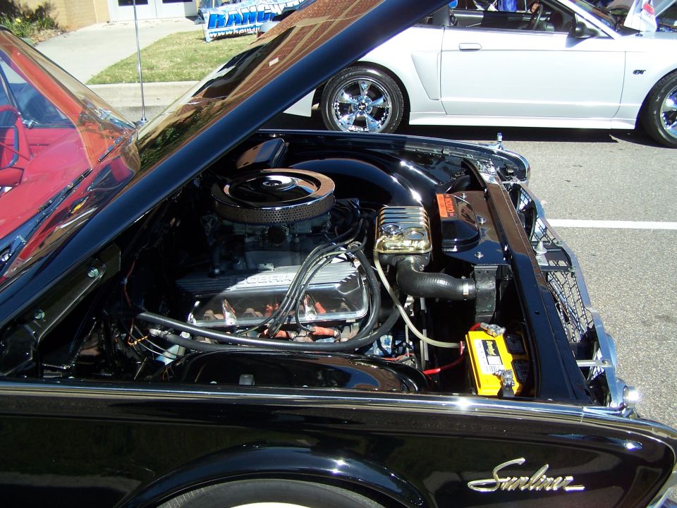Black car engine bay with chrome accents and a yellow battery. A silver convertible is visible behind