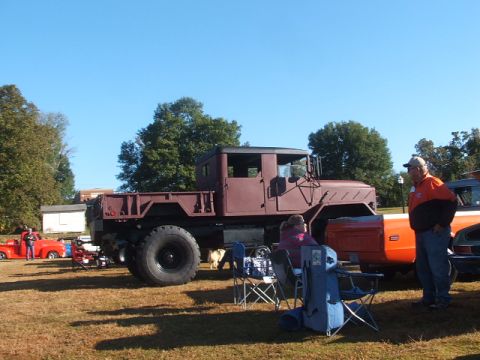1971 Ford Ranchero - A large maroon military truck is parked on a grassy field with people gathered around, alongside other vehicles under a clear blue sky.