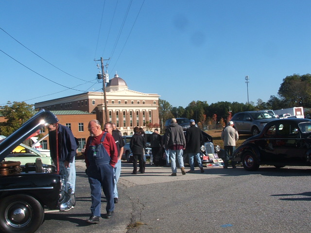 Black classic car with hood open, people gathered around, building in background