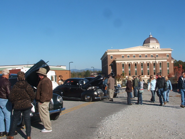 Black coupe with hood open, parked on a street with people around. Building in background