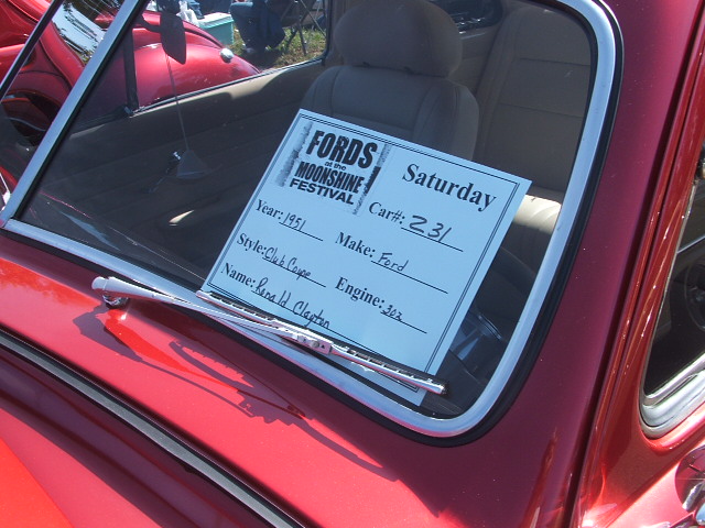 Interior of a red Ford car with a sign that reads 'FORDS at the MOONSHINE FESTIVAL Saturday Car#: 231 Year: 1951 Make: Ford Style: Club Coupe Engine: 302 Name: Randal H. Clayton'