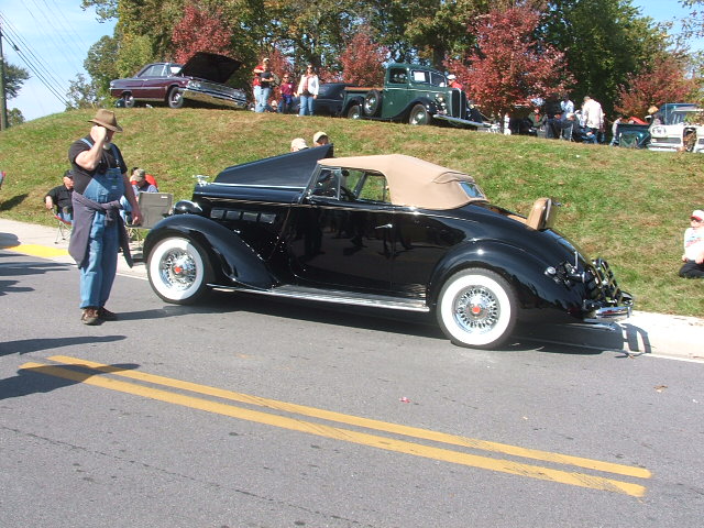 Black convertible with tan top and whitewall tires parked on a street, man in overalls standing nearby. Other cars and people on grassy hill in background