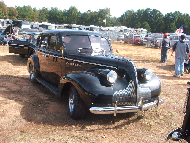 Black vintage sedan with chrome grille and headlights parked on dirt lot with RVs and people in background