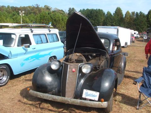 1971 Ford Ranchero - A black vintage car with its hood open is parked next to a light blue van with surfboards on its roof.