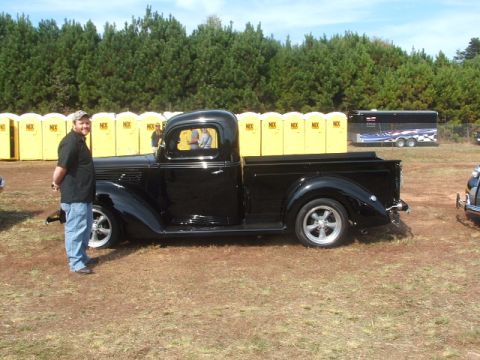 1971 Ford Ranchero - A man poses with a shiny black vintage pickup truck on a grassy field with portable toilets and a trailer behind it tow.