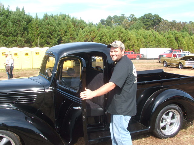 Black pickup truck, driver's side door open, man in camouflage hat smiling, chrome grille, custom wheels