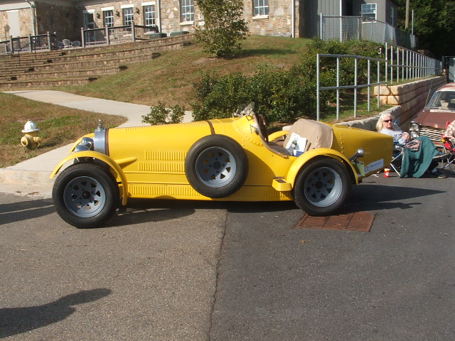 Yellow vintage-style open-top car with a side-mounted spare tire and a tan convertible top, parked on a street