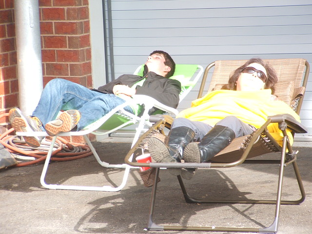 Two people relax in lounge chairs outdoors, with a brick wall and garage door in the background