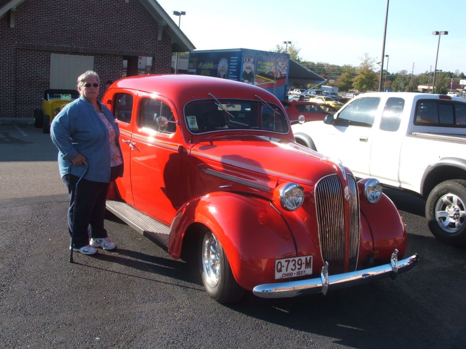 Red 1937 Plymouth sedan with a woman standing beside it. White pickup truck parked to the right. License plate reads Q-739-M