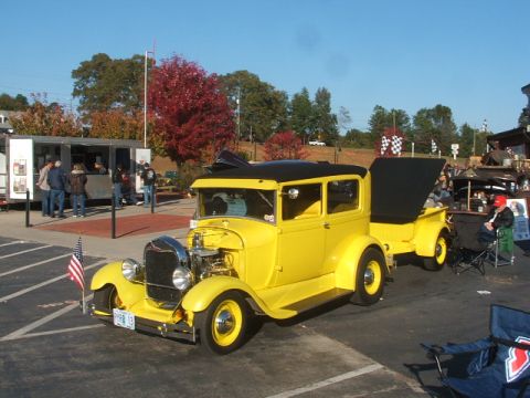 1971 Ford Ranchero - A yellow 1928 Ford Model A sedan is parked with a trailer, with people near a food truck in the background.
