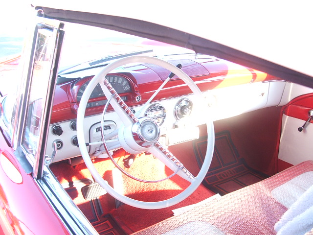 Interior view of a classic car, showing a white steering wheel, red dashboard, and floor mats. The year appears to be 1955