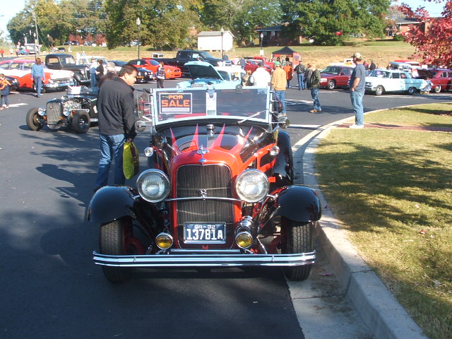 Front view of a red and black hot rod with flames, "FOR SALE" sign on windshield, at a car show
