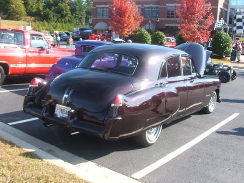 1971 Ford Ranchero - A dark purple sedan is parked in a lot with other vehicles, including a red truck and a hot rod.