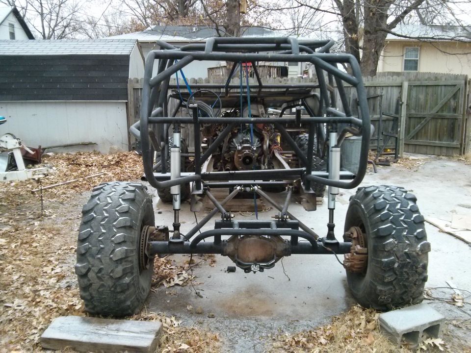 Off-road buggy frame with large tires, exposed engine, and roll cage. Rear view