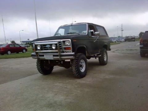1986 Ford Bronco - This lifted Ford Bronco is parked on a concrete surface with other vehicles in the background.
