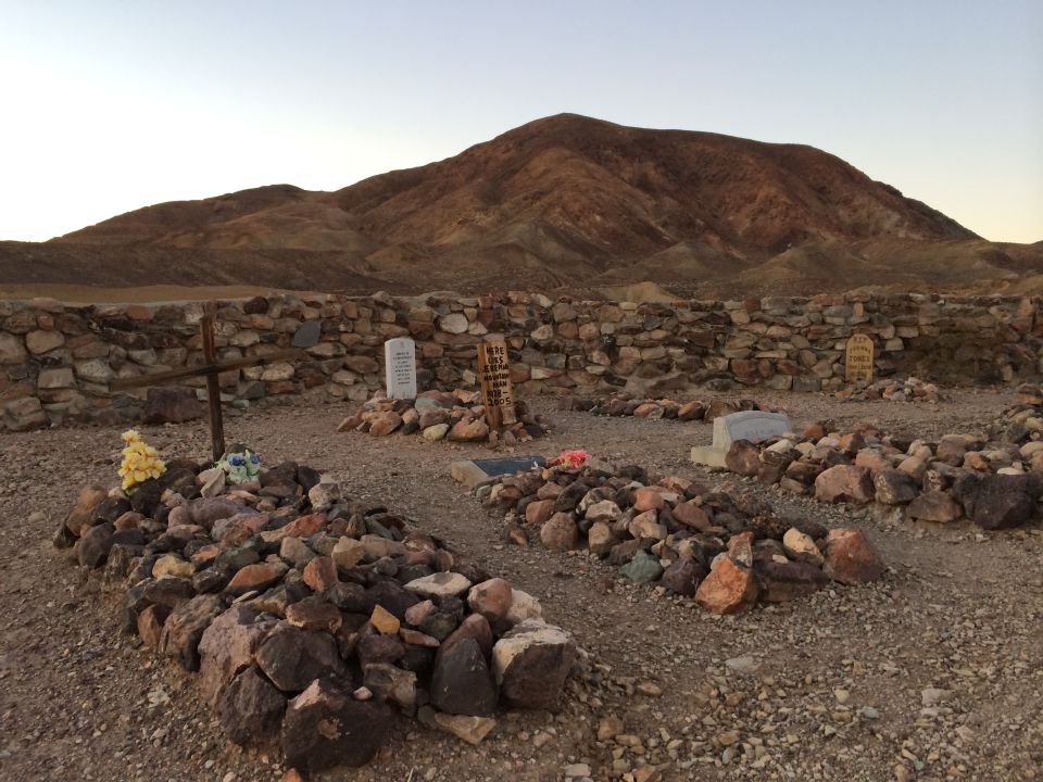 Desert cemetery with stone walls, graves marked by rocks and crosses, and a mountain range under a clear sky