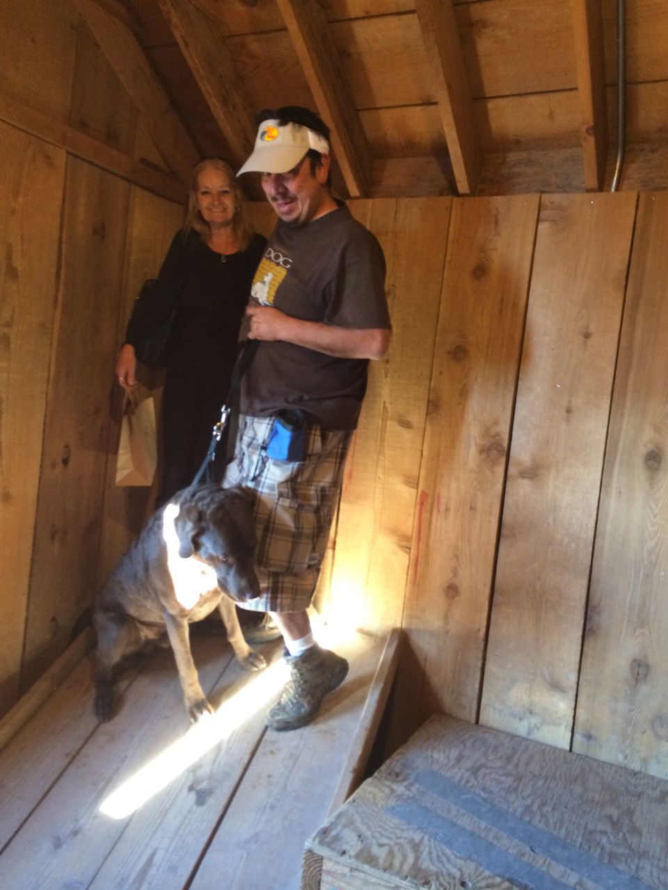 Man, woman, and dog stand inside a rustic wooden room. Man wears a visor and plaid shorts