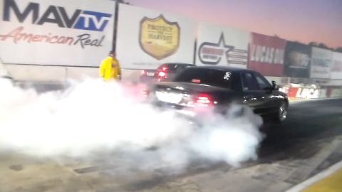 2003 Ford Crown Victoria - black full-size sedan performing a smoky burnout at a drag strip lined with MavTV and sponsor banners.