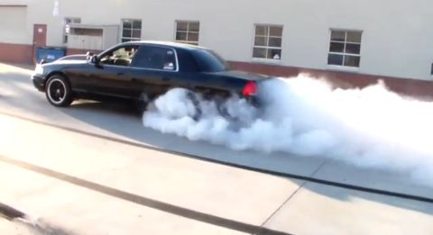 2003 Ford Crown Victoria - black full-size sedan doing a burnout in a parking lot, producing a large cloud of tire smoke.