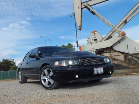 2003 Ford Crown Victoria - black Ford Crown Victoria with halo headlights and 'EVIL VIC' plate parked on gravel beside an oil pumpjack.