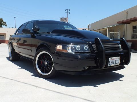 2003 Ford Crown Victoria - black Ford Crown Victoria with a push bar, halo headlights, and 'EVIL VIC' plate parked in an industrial lot.