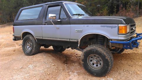 1990 Ford Bronco - Modified 1990 Ford Bronco with off-road tires and custom bumper sits on a dirt trail.
