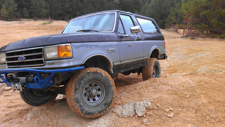 Gray and black Ford Bronco with a blue aftermarket bumper and winch, stuck in mud on a dirt trail