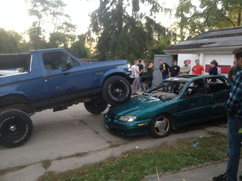 1996 Ford Bronco - A blue Ford Bronco is parked on top of a wrecked green sedan, with a group of onlookers.