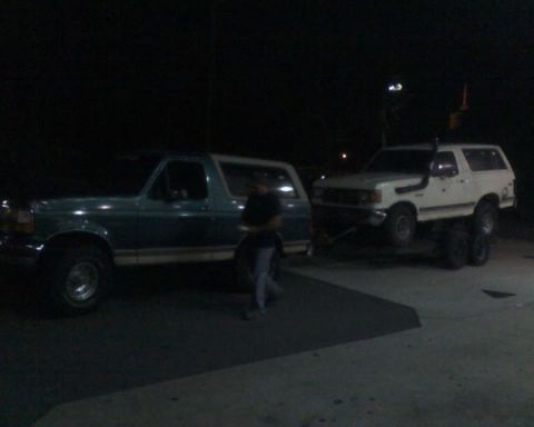 1996 Ford Bronco - A green 1996 Ford Bronco tows a white Ford Bronco, both seen from the side in the dark.