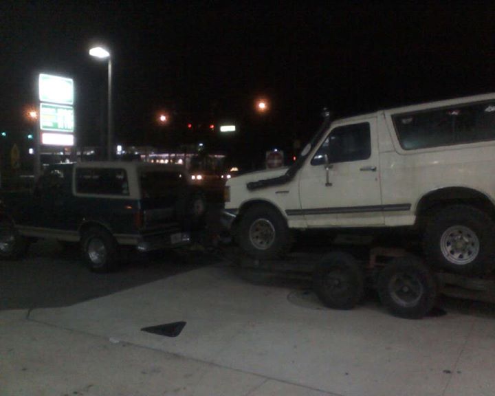 Dark blue Ford Bronco parked next to a white Ford Bronco on a trailer at night