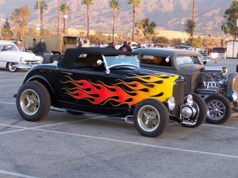 1990 Ford Bronco - A black hot rod with flame graphics is parked next to a grey hot rod, with palm trees and mountains in the background.