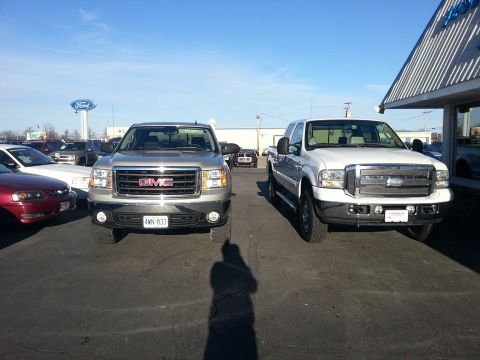 1995 Ford F-150 - A silver GMC pickup truck is parked next to a white Ford pickup truck under a bright blue sky.