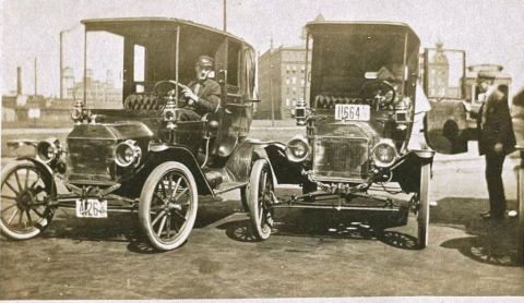 1912 Ford Model T - Two vintage automobiles are parked on a street, with one driver visible and another man standing near the second vehicle.
