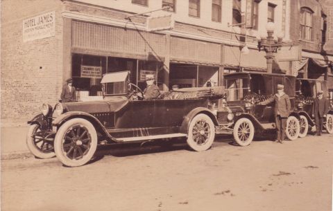 1914 Ford Model T - A line of vintage automobiles is parked along the curb in front of businesses, including the Hotel James and Columbus Cafe.