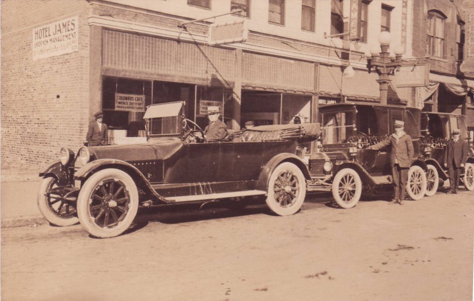 Several old touring cars parked in a row on a street in front of buildings. One car has people inside