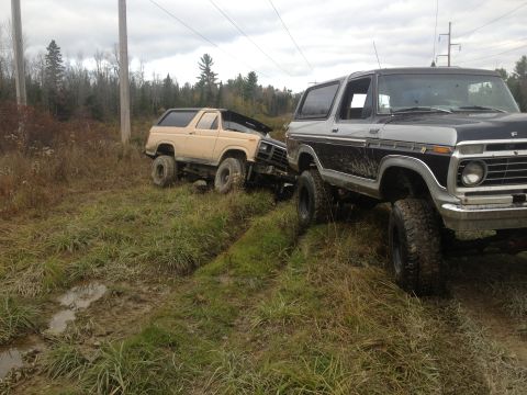 1970 Ford F-250 - Two Ford Broncos are stuck in a muddy, grassy area with trees and power lines in the background.