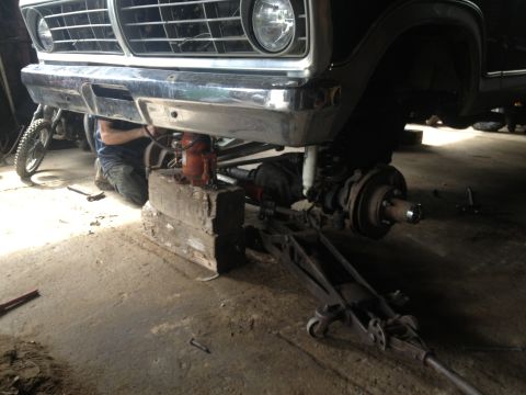 1970 Ford F-250 - A mechanic works on the front suspension of a black Ford F-Series truck, which is elevated on blocks and a jack.