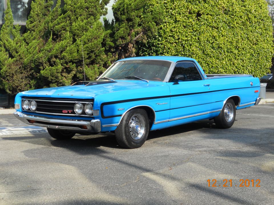 Bright blue Ford Ranchero GT with black stripes and hood scoop, parked in front of tall hedges