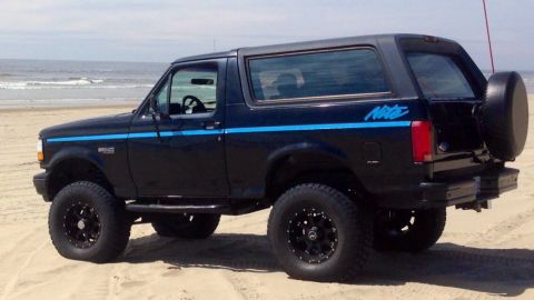 1992 Ford Bronco - A black 1992 Ford Bronco with blue racing stripes is parked on a beach near the ocean.