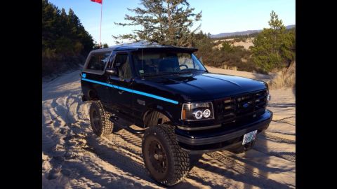 1992 Ford Bronco - A black 1992 Ford Bronco with a blue stripe drives on a sandy path, with trees and dunes in the background.