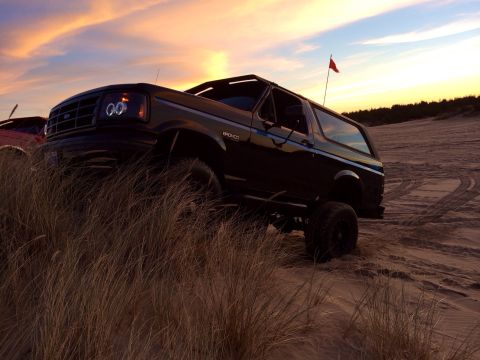1992 Ford Bronco - A black 1992 Ford Bronco navigates sand dunes under a colorful sunset sky.