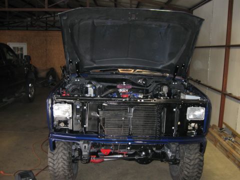 1982 Ford Bronco - Front view of a blue truck with its hood open, revealing the engine and grille area. Another vehicle is partially visible to the left.