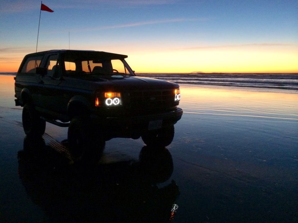 1992 Ford Bronco on wet sand with ocean waves and sunset in background