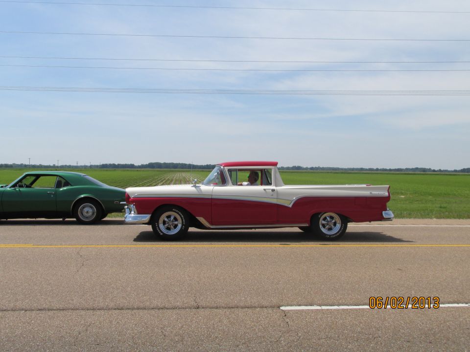 Red and white 1957 Ford Ranchero in side view on a rural road with a green car behind it