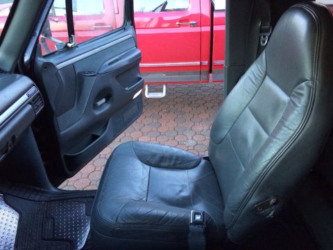 1992 Ford Bronco - The interior of a vehicle shows a black leather seat and an open door revealing a red pickup truck outside.